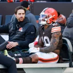Ein Footballspieler der Browns, der Quinshon Judkins ähnelt und die Uniformnummer 10 trägt, sitzt auf einem Krankenwagen, neben ihm die Trainer. Sein linkes Bein ist gestreckt und gewickelt, was auf eine Beinverletzung während eines Spiels an der kalten Seitenlinie hinweist. Diese Beschreibung wurde mit der FootballR KI automatisch generiert.
