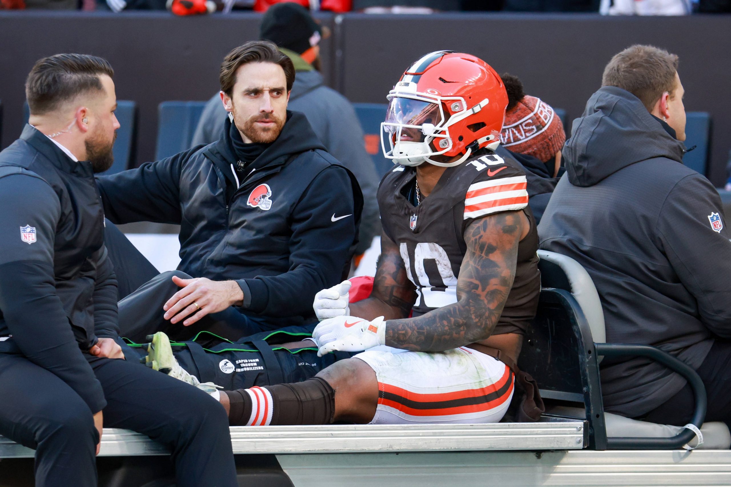 Ein Footballspieler der Browns, der Quinshon Judkins ähnelt und die Uniformnummer 10 trägt, sitzt auf einem Krankenwagen, neben ihm die Trainer. Sein linkes Bein ist gestreckt und gewickelt, was auf eine Beinverletzung während eines Spiels an der kalten Seitenlinie hinweist. Diese Beschreibung wurde mit der FootballR KI automatisch generiert.
