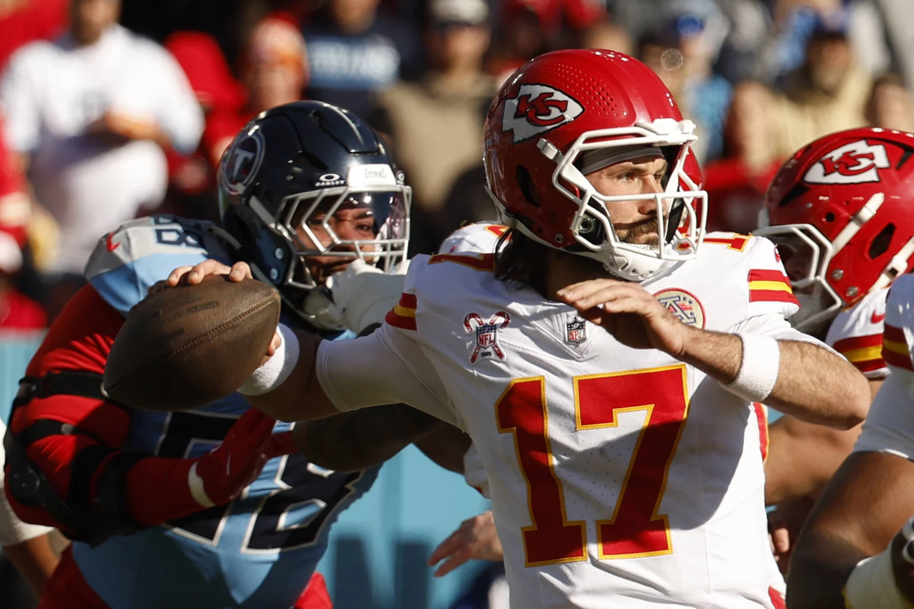 Gardner Minshew - Ein Quarterback der Kansas City Chiefs (#17) bereitet sich darauf vor, einen Football zu werfen, während er einem Verteidiger der Tennessee Titans (#98) während eines NFL-Spiels ausweicht. Der Chiefs-Quarterback trägt einen roten Helm und ein weißes Trikot; der Verteidiger trägt einen dunklen Helm und ein hellblaues Trikot. Diese Beschreibung wurde mit der FootballR KI automatisch generiert.