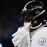DK Metcalf - Ein Footballspieler mit schwarzem Helm und weißer Steelers-Uniform, auf deren Trikot das Logo der Mannschaft zu sehen ist, hält sich die behandschuhten Hände vors Gesicht. Der Hintergrund ist unscharf mit dunkelblauen Tönen und fängt den Geist der Herausforderung ein. Diese Beschreibung wurde mit der FootballR KI automatisch generiert.