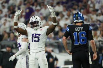 Ein Footballspieler in einem weiß-lila Trikot reißt auf dem Spielfeld feierlich beide Arme hoch. In der Nähe steht Detroit Lions Quarterback Goff mit der Nummer 16 mit dem Rücken zu den Fans, die das Stadion während der intensiven Playoff-Atmosphäre füllen. Diese Beschreibung wurde mit der FootballR KI automatisch generiert.