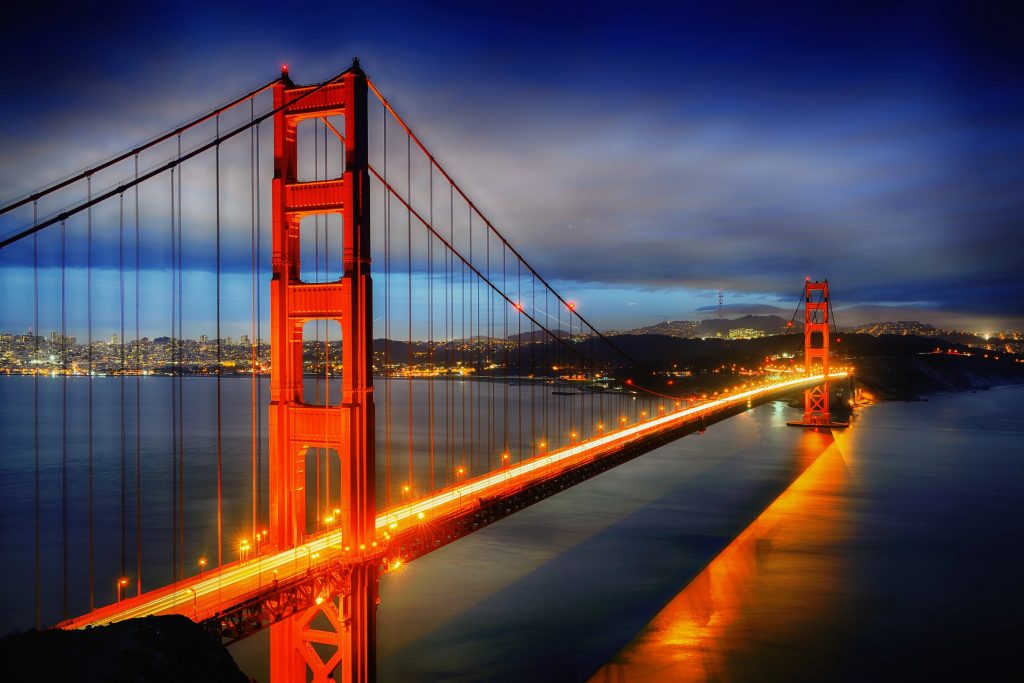 Die Golden Gate Bridge leuchtet in der Abenddämmerung in orangefarbenem Licht und überspannt das ruhige blaue Wasser in San Francisco. Die beleuchtete Brücke kontrastiert mit dem dunklen Himmel und den Lichtern der Stadt und schafft eine dramatische Szenerie, die sich perfekt für jeden Super Bowl 2026 Event-Guide eignet. Diese Beschreibung wurde mit der FootballR KI automatisch generiert.