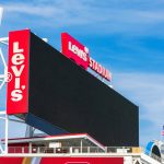 Eine große, schwarze, leere elektronische Anzeigetafel im Levi's Stadium vor einem blauen Himmel. Hohe rote Tafeln auf der linken und oberen Seite zeigen das weiße Logo des Levi's® Stadium. Das Stadion, in dem der Super Bowl LX ausgetragen wird, zeigt unten rechts eine amerikanische Flagge und darunter Dachstrukturen. Diese Beschreibung wurde mit der FootballR KI automatisch generiert.