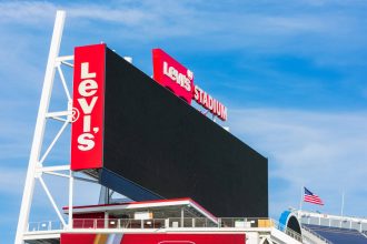 Eine große, schwarze, leere elektronische Anzeigetafel im Levi's Stadium vor einem blauen Himmel. Hohe rote Tafeln auf der linken und oberen Seite zeigen das weiße Logo des Levi's® Stadium. Das Stadion, in dem der Super Bowl LX ausgetragen wird, zeigt unten rechts eine amerikanische Flagge und darunter Dachstrukturen. Diese Beschreibung wurde mit der FootballR KI automatisch generiert.