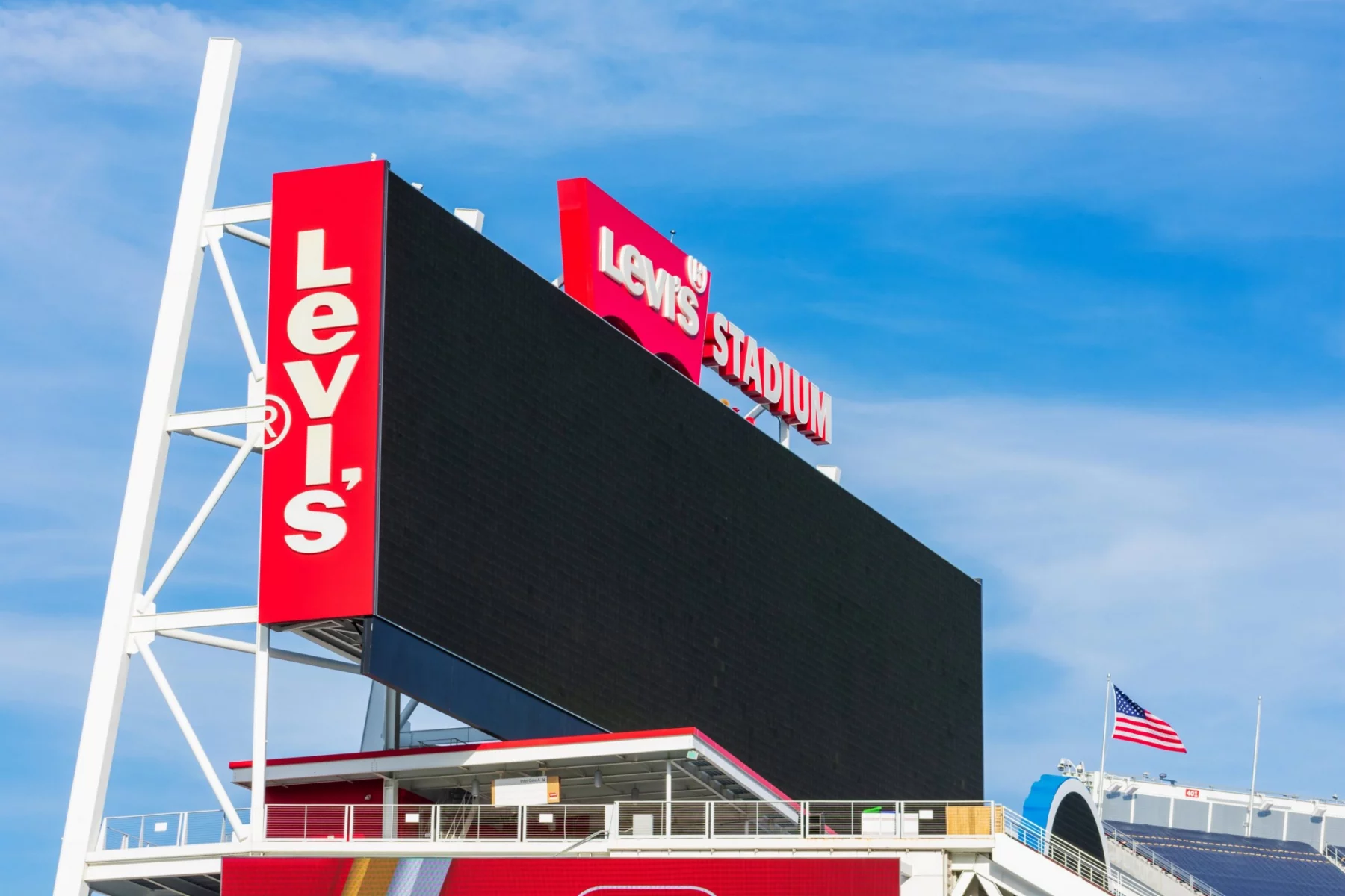 Eine große, schwarze, leere elektronische Anzeigetafel im Levi's Stadium vor einem blauen Himmel. Hohe rote Tafeln auf der linken und oberen Seite zeigen das weiße Logo des Levi's® Stadium. Das Stadion, in dem der Super Bowl LX ausgetragen wird, zeigt unten rechts eine amerikanische Flagge und darunter Dachstrukturen. Diese Beschreibung wurde mit der FootballR KI automatisch generiert.