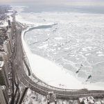 Sean McVay übers Wetter: Luftaufnahme der verschneiten Uferlinie von Chicago-Spiel und eines zugefrorenen Sees mit rissigen Eismustern. Schneebedeckte Straßen schlängeln sich links an Hochhäusern vorbei. Die winterliche, bedeckte Szene strahlt Kälte aus, mit grauem Himmel und wenig Verkehr. Diese Beschreibung wurde mit der FootballR KI automatisch generiert.