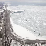 Sean McVay übers Wetter: Luftaufnahme der verschneiten Uferlinie von Chicago-Spiel und eines zugefrorenen Sees mit rissigen Eismustern. Schneebedeckte Straßen schlängeln sich links an Hochhäusern vorbei. Die winterliche, bedeckte Szene strahlt Kälte aus, mit grauem Himmel und wenig Verkehr. Diese Beschreibung wurde mit der FootballR KI automatisch generiert.