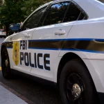 Ein weißes FBI-Polizeiauto parkt auf einer Straße in der Stadt und trägt die Aufschrift "FBI POLICE" und ein Abzeichensymbol. Orangefarbene Verkehrskegel säumen die Straße, während im Hintergrund Bäume und Gebäude zu sehen sind - keine Spur von Schmerzmitteln oder Jim Irsay in der Nähe. Diese Beschreibung wurde mit der FootballR KI automatisch generiert.