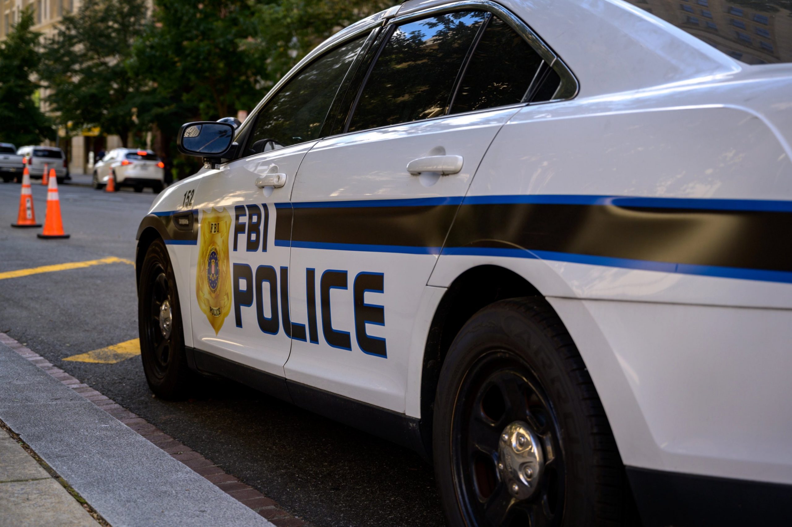 Ein weißes FBI-Polizeiauto parkt auf einer Straße in der Stadt und trägt die Aufschrift "FBI POLICE" und ein Abzeichensymbol. Orangefarbene Verkehrskegel säumen die Straße, während im Hintergrund Bäume und Gebäude zu sehen sind - keine Spur von Schmerzmitteln oder Jim Irsay in der Nähe. Diese Beschreibung wurde mit der FootballR KI automatisch generiert.