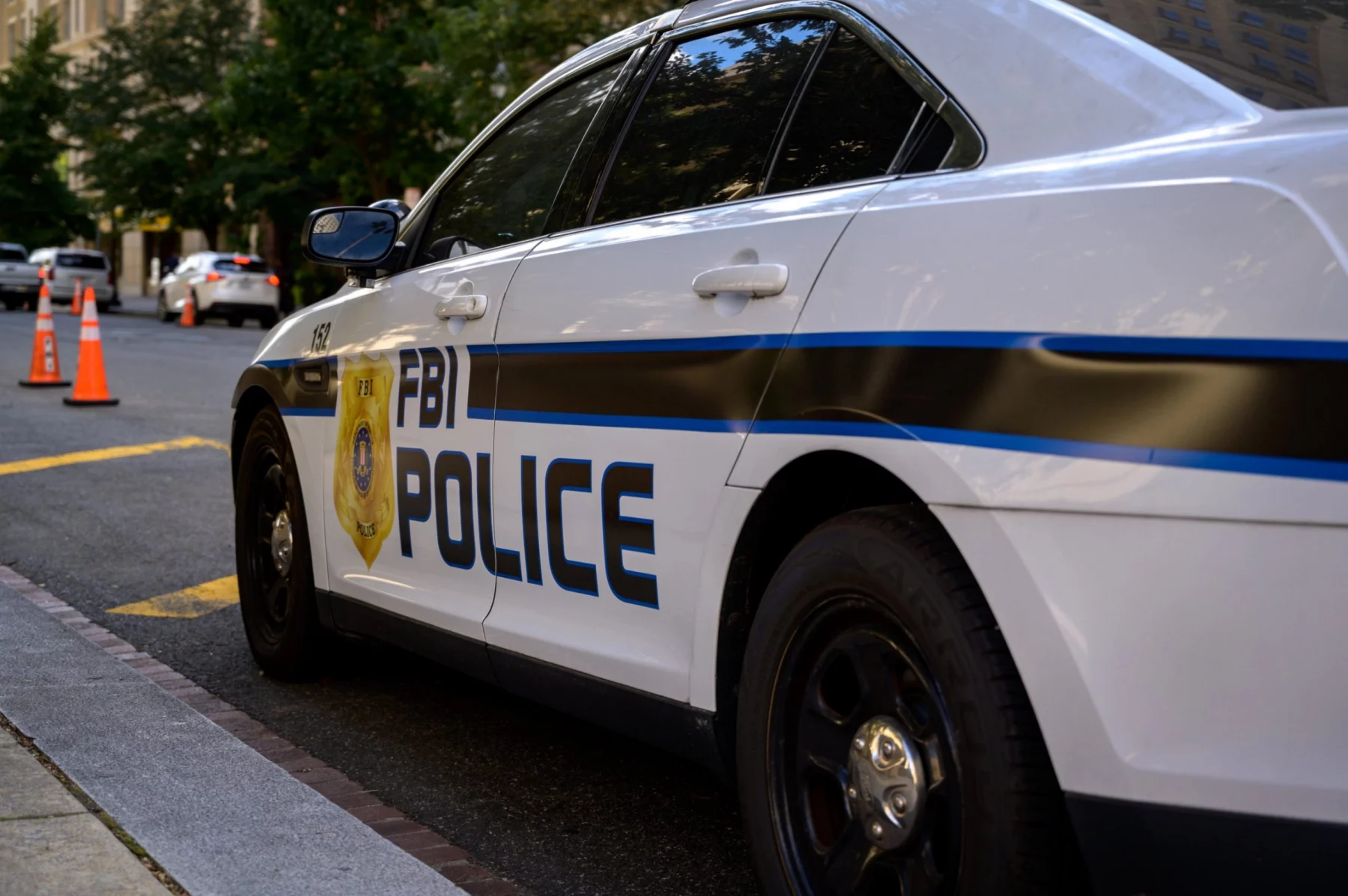 Ein weißes FBI-Polizeiauto parkt auf einer Straße in der Stadt und trägt die Aufschrift "FBI POLICE" und ein Abzeichensymbol. Orangefarbene Verkehrskegel säumen die Straße, während im Hintergrund Bäume und Gebäude zu sehen sind - keine Spur von Schmerzmitteln oder Jim Irsay in der Nähe. Diese Beschreibung wurde mit der FootballR KI automatisch generiert.