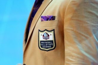 Nahaufnahme einer hellbraunen Anzugsjacke mit dem Aufnäher Pro Football Hall of Fame Enshrinee. Die prestigeträchtige Aufnahme in die Hall of Fame durch das Auswahlkomitee wird durch ein violettes und goldenes Einstecktuch vor hellblauem Hintergrund hervorgehoben. Diese Beschreibung wurde mit der FootballR KI automatisch generiert.