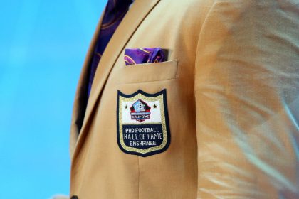 Nahaufnahme einer hellbraunen Anzugsjacke mit dem Aufnäher Pro Football Hall of Fame Enshrinee. Die prestigeträchtige Aufnahme in die Hall of Fame durch das Auswahlkomitee wird durch ein violettes und goldenes Einstecktuch vor hellblauem Hintergrund hervorgehoben. Diese Beschreibung wurde mit der FootballR KI automatisch generiert.