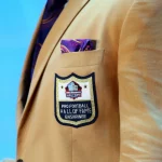 Nahaufnahme einer hellbraunen Anzugsjacke mit dem Aufnäher Pro Football Hall of Fame Enshrinee. Die prestigeträchtige Aufnahme in die Hall of Fame durch das Auswahlkomitee wird durch ein violettes und goldenes Einstecktuch vor hellblauem Hintergrund hervorgehoben. Diese Beschreibung wurde mit der FootballR KI automatisch generiert.