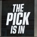 In großen, fetten weißen Buchstaben steht auf einem Bildschirm "THE PICK IS IN". Der dunkle Hintergrund und die dramatische Atmosphäre erinnern an die Spannung des NFL Draft 2026, bei dem die Teams auf die Bekanntgabe der Reihenfolge und des Nr.-1-Picks warten. Diese Beschreibung wurde mit der FootballR KI automatisch generiert.