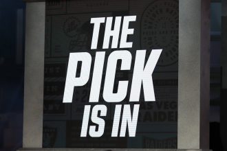In großen, fetten weißen Buchstaben steht auf einem Bildschirm "THE PICK IS IN". Der dunkle Hintergrund und die dramatische Atmosphäre erinnern an die Spannung des NFL Draft 2026, bei dem die Teams auf die Bekanntgabe der Reihenfolge und des Nr.-1-Picks warten. Diese Beschreibung wurde mit der FootballR KI automatisch generiert.