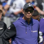 Todd Monken trägt eine Sonnenbrille, eine schwarze Ravens-Kappe und einen lilafarbenen Team-Kapuzenpulli und steht während eines sonnigen Footballspiels an der Seitenlinie. Andere Mitarbeiter und Spieler in Ravens-Kleidung sind im Hintergrund zu sehen. Diese Beschreibung wurde mit der FootballR KI automatisch generiert.