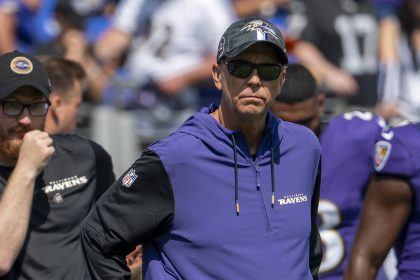 Todd Monken trägt eine Sonnenbrille, eine schwarze Ravens-Kappe und einen lilafarbenen Team-Kapuzenpulli und steht während eines sonnigen Footballspiels an der Seitenlinie. Andere Mitarbeiter und Spieler in Ravens-Kleidung sind im Hintergrund zu sehen. Diese Beschreibung wurde mit der FootballR KI automatisch generiert.
