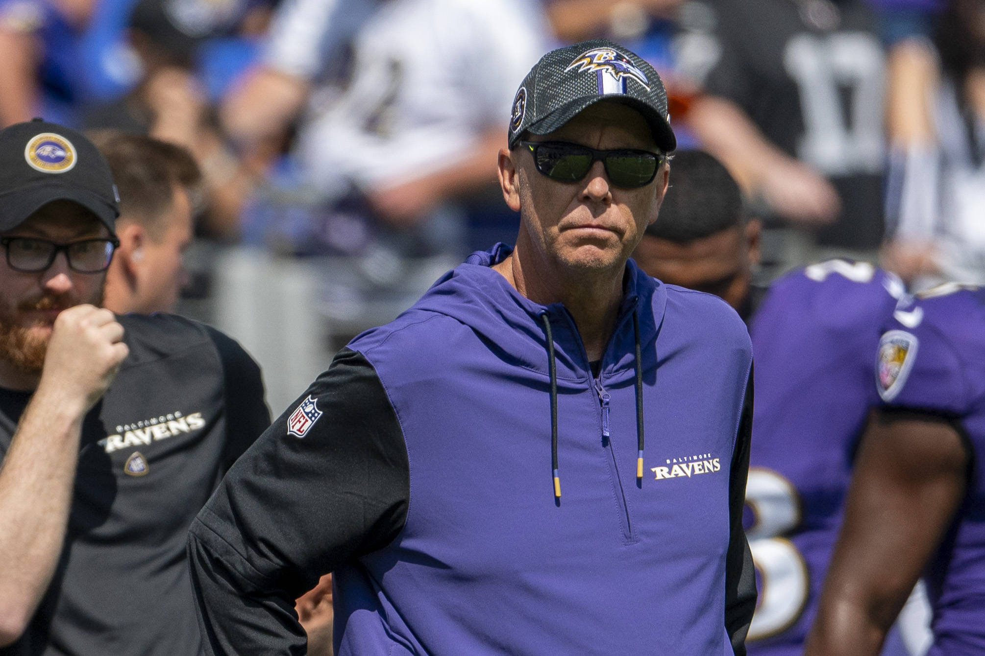 Todd Monken trägt eine Sonnenbrille, eine schwarze Ravens-Kappe und einen lilafarbenen Team-Kapuzenpulli und steht während eines sonnigen Footballspiels an der Seitenlinie. Andere Mitarbeiter und Spieler in Ravens-Kleidung sind im Hintergrund zu sehen. Diese Beschreibung wurde mit der FootballR KI automatisch generiert.