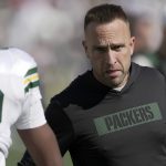 Jeff Hafley - Ein Mann mit einem schwarzen "PACKERS"-Kapuzenpulli, möglicherweise der Cheftrainer, blickt aufmerksam auf einen Footballspieler in einer Green Bay Packers-Uniform auf einem sonnigen Feld. Der Rücken des Spielers ist sichtbar und zeigt teilweise seinen Namen und seine Nummer während des Trainings oder eines Spiels. Diese Beschreibung wurde mit der FootballR KI automatisch generiert.