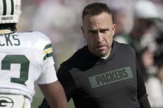 Jeff Hafley - Ein Mann mit einem schwarzen "PACKERS"-Kapuzenpulli, möglicherweise der Cheftrainer, blickt aufmerksam auf einen Footballspieler in einer Green Bay Packers-Uniform auf einem sonnigen Feld. Der Rücken des Spielers ist sichtbar und zeigt teilweise seinen Namen und seine Nummer während des Trainings oder eines Spiels. Diese Beschreibung wurde mit der FootballR KI automatisch generiert.