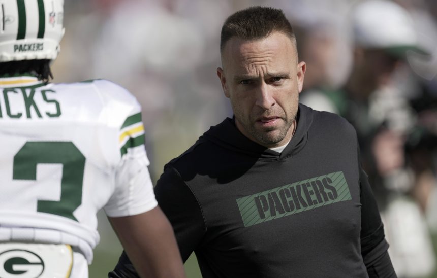 Jeff Hafley - Ein Mann mit einem schwarzen "PACKERS"-Kapuzenpulli, möglicherweise der Cheftrainer, blickt aufmerksam auf einen Footballspieler in einer Green Bay Packers-Uniform auf einem sonnigen Feld. Der Rücken des Spielers ist sichtbar und zeigt teilweise seinen Namen und seine Nummer während des Trainings oder eines Spiels. Diese Beschreibung wurde mit der FootballR KI automatisch generiert.