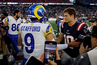 MVP - Ein Rams-Spieler in einem blau-gelben Matthew STAFFORD 9-Trikot schüttelt Drake Maye, der die Nummer 10 der Patriots trägt, auf dem Football-Feld die Hand - ein Moment zwischen den NFL-Award-Finalisten inmitten von Spielern, Medien, Fans und einer Stadionkulisse. Diese Beschreibung wurde mit der FootballR KI automatisch generiert.