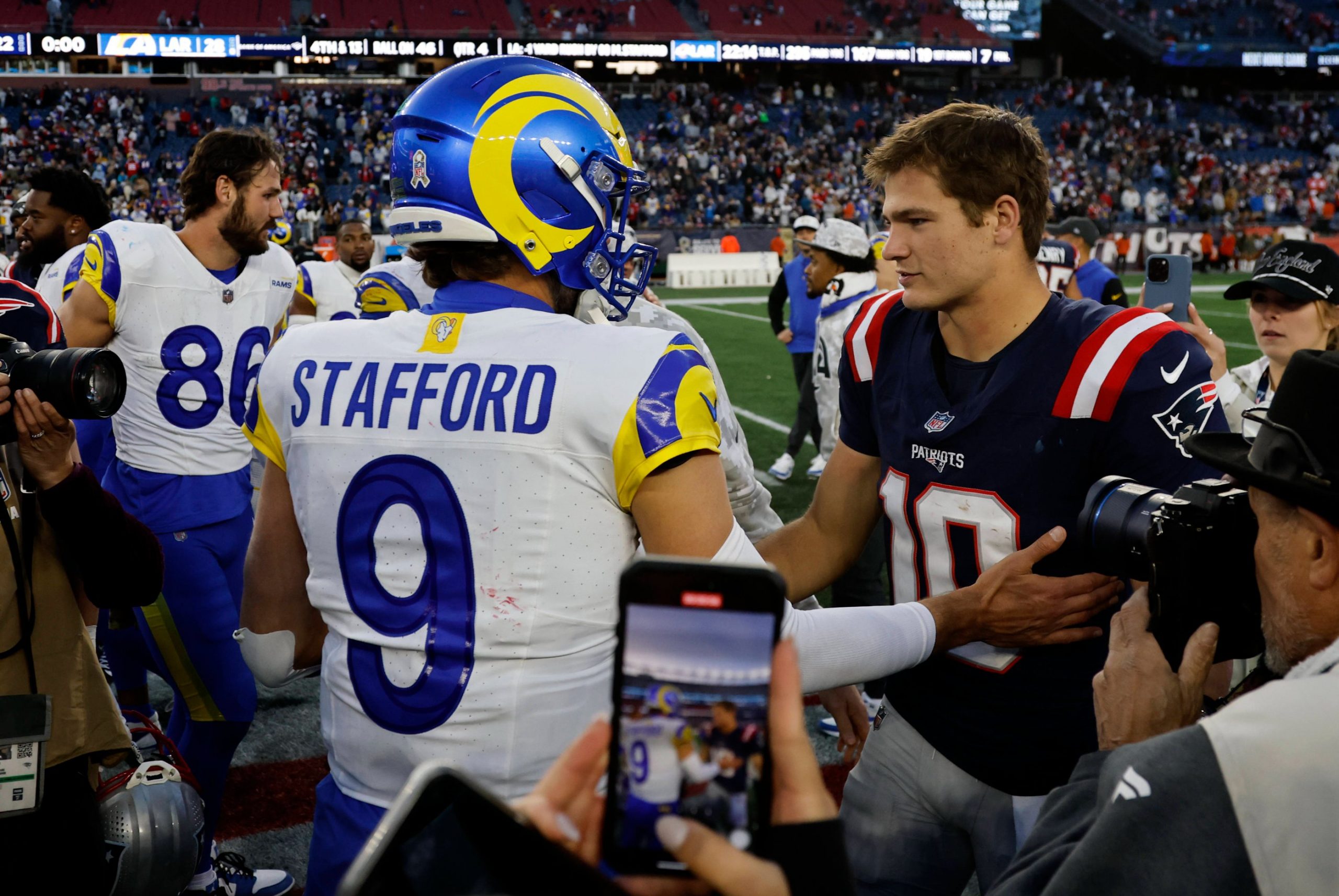 MVP - Ein Rams-Spieler in einem blau-gelben Matthew STAFFORD 9-Trikot schüttelt Drake Maye, der die Nummer 10 der Patriots trägt, auf dem Football-Feld die Hand - ein Moment zwischen den NFL-Award-Finalisten inmitten von Spielern, Medien, Fans und einer Stadionkulisse. Diese Beschreibung wurde mit der FootballR KI automatisch generiert.