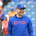 Ein lächelnder Mann mit blauem "BILLS"-Kapuzenpulli und passender Mütze steht in einem Stadion mit leeren blauen Sitzen und freut sich möglicherweise darauf, Head Coach Joe Brady zu sehen. Eine weitere Person in einem grauen Sweatshirt steht neben ihm, weitere Personen sind im Hintergrund zu sehen. Diese Beschreibung wurde mit der FootballR KI automatisch generiert.