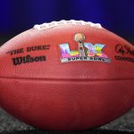 Eine Nahaufnahme eines offiziellen Super Bowl LX Footballs liegt auf einer dunklen, strukturierten Oberfläche. Der Ball zeigt das Super Bowl LX Logo, eine silberne Trophäe, ein farbenfrohes Stadtbild, schwarze Unterschriften, das "The Duke"-Branding von Wilson und eine subtile Anspielung auf die Super Bowl LX-Wertung. Diese Beschreibung wurde mit der FootballR KI automatisch generiert.