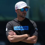 Jesse Minter - Ein Mann mit weißer Mütze, verspiegelter Sonnenbrille und schwarzem Kapuzenpulli mit blauem "CHARGERS"-Logo steht mit verschränkten Armen im Freien und wirkt konzentriert - wie Cheftrainer Jesse Minter während der Trainingseinheiten der Baltimore Ravens. Diese Beschreibung wurde mit der FootballR KI automatisch generiert.
