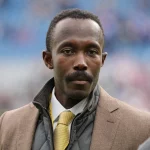 Ein Mann mit kurzen Haaren und Schnurrbart, der einen braunen Mantel über einer grauen Steppjacke und eine gelbe Krawatte trägt, steht draußen neben den verschwommenen blauen Stadionsitzen. Er hat einen ernsten Gesichtsausdruck - es ist der Vikings-Geschäftsführer Kwesi Adofo-Mensah. Diese Beschreibung wurde mit der FootballR KI automatisch generiert.