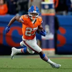 J.K. Dobbins - Ein Footballspieler in einer orange-blauen Broncos-Uniform mit der Nummer 27 läuft mit dem Ball auf einem nächtlichen Spielfeld während des AFC Championship Game. Er trägt einen Helm, Handschuhe und weiße Stollenschuhe. Im unscharfen Hintergrund sind Fans und eine blaue Broncos-Fahne zu sehen. Diese Beschreibung wurde mit der FootballR KI automatisch generiert.