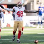 Tatum Bethune - Ein Abwehrspieler der San Francisco 49ers mit der Trikotnummer 48 breitet seine Arme auf dem Spielfeld aus, während vor ihm ein Football auf dem Boden liegt. Er trägt ein weißes Trikot, eine goldene Hose und einen Helm und sticht als Leistungsträger zwischen den verschwommenen Gegnern im Hintergrund hervor. Diese Beschreibung wurde mit der FootballR KI automatisch generiert.