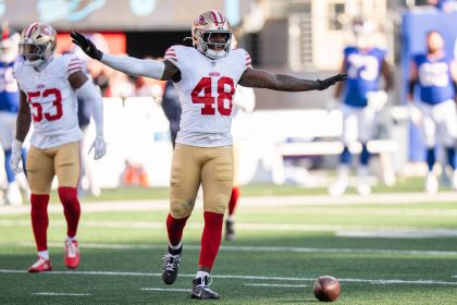 Tatum Bethune - Ein Abwehrspieler der San Francisco 49ers mit der Trikotnummer 48 breitet seine Arme auf dem Spielfeld aus, während vor ihm ein Football auf dem Boden liegt. Er trägt ein weißes Trikot, eine goldene Hose und einen Helm und sticht als Leistungsträger zwischen den verschwommenen Gegnern im Hintergrund hervor. Diese Beschreibung wurde mit der FootballR KI automatisch generiert.