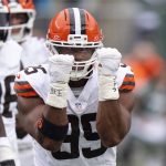Ein Football-Spieler in einer weißen Cleveland Browns-Uniform und einem orangefarbenen Helm, wahrscheinlich Myles Garrett, hebt beide behandschuhten Fäuste vor sein Gesicht und feiert einen NFL-Moment. Andere Spieler in ähnlichen Uniformen sind auf dem grünen Football-Feld zu sehen. Diese Beschreibung wurde mit der FootballR KI automatisch generiert.