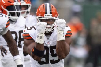 Ein Football-Spieler in einer weißen Cleveland Browns-Uniform und einem orangefarbenen Helm, wahrscheinlich Myles Garrett, hebt beide behandschuhten Fäuste vor sein Gesicht und feiert einen NFL-Moment. Andere Spieler in ähnlichen Uniformen sind auf dem grünen Football-Feld zu sehen. Diese Beschreibung wurde mit der FootballR KI automatisch generiert.