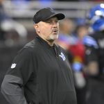 Der Offensive Coordinator der Detroit Lions, John Morton, trägt eine schwarze Jacke und eine Kappe mit dem Teamlogo und steht auf dem Spielfeld. Konzentriert und aufmerksam beobachtet er, wie sich ein Spieler in einer blau-schwarzen Uniform im verschwommenen Hintergrund bewegt. Diese Beschreibung wurde mit der FootballR KI automatisch generiert.
