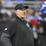 Der Offensive Coordinator der Detroit Lions, John Morton, trägt eine schwarze Jacke und eine Kappe mit dem Teamlogo und steht auf dem Spielfeld. Konzentriert und aufmerksam beobachtet er, wie sich ein Spieler in einer blau-schwarzen Uniform im verschwommenen Hintergrund bewegt. Diese Beschreibung wurde mit der FootballR KI automatisch generiert.