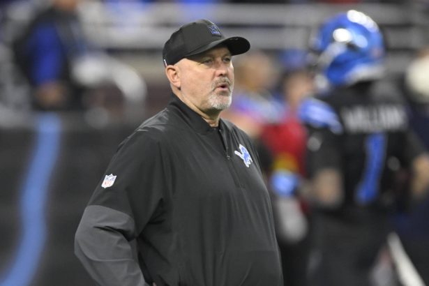 Der Offensive Coordinator der Detroit Lions, John Morton, trägt eine schwarze Jacke und eine Kappe mit dem Teamlogo und steht auf dem Spielfeld. Konzentriert und aufmerksam beobachtet er, wie sich ein Spieler in einer blau-schwarzen Uniform im verschwommenen Hintergrund bewegt. Diese Beschreibung wurde mit der FootballR KI automatisch generiert.