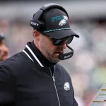 Kevin Patullo, Offensive Coordinator der Philadelphia Eagles, trägt eine Sonnenbrille, eine schwarze "OPPORTUNITY"-Mütze und ein Headset und blickt auf seinen Spielplan. Er trägt eine schwarze Jacke mit weißen Streifen und einem Eagles-Logo inmitten verschwommener Stadionsitze. Diese Beschreibung wurde mit der FootballR KI automatisch generiert.