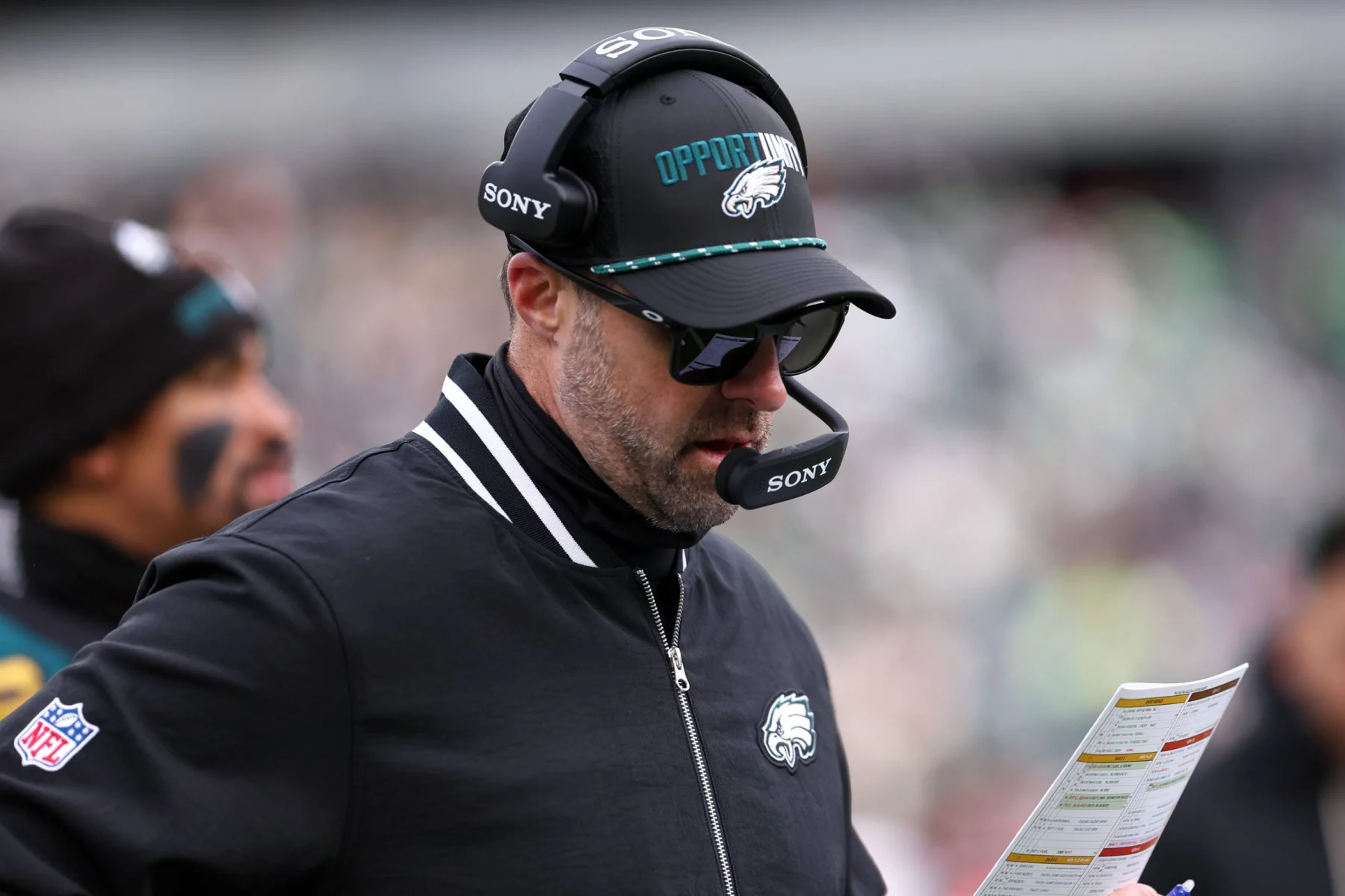 Kevin Patullo, Offensive Coordinator der Philadelphia Eagles, trägt eine Sonnenbrille, eine schwarze "OPPORTUNITY"-Mütze und ein Headset und blickt auf seinen Spielplan. Er trägt eine schwarze Jacke mit weißen Streifen und einem Eagles-Logo inmitten verschwommener Stadionsitze. Diese Beschreibung wurde mit der FootballR KI automatisch generiert.