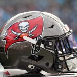 Fünf Trainer mussten gehen - Eine Nahaufnahme eines Footballhelms der Tampa Bay Buccaneers zeigt ein silbernes und rotes Design mit dem ikonischen Piratenflaggen-Logo. Der Helm, der mit Cheftrainer Bowles verbunden ist, ruht auf einer Fläche mit einem unscharfen Stadionhintergrund. Diese Beschreibung wurde mit der FootballR KI automatisch generiert.