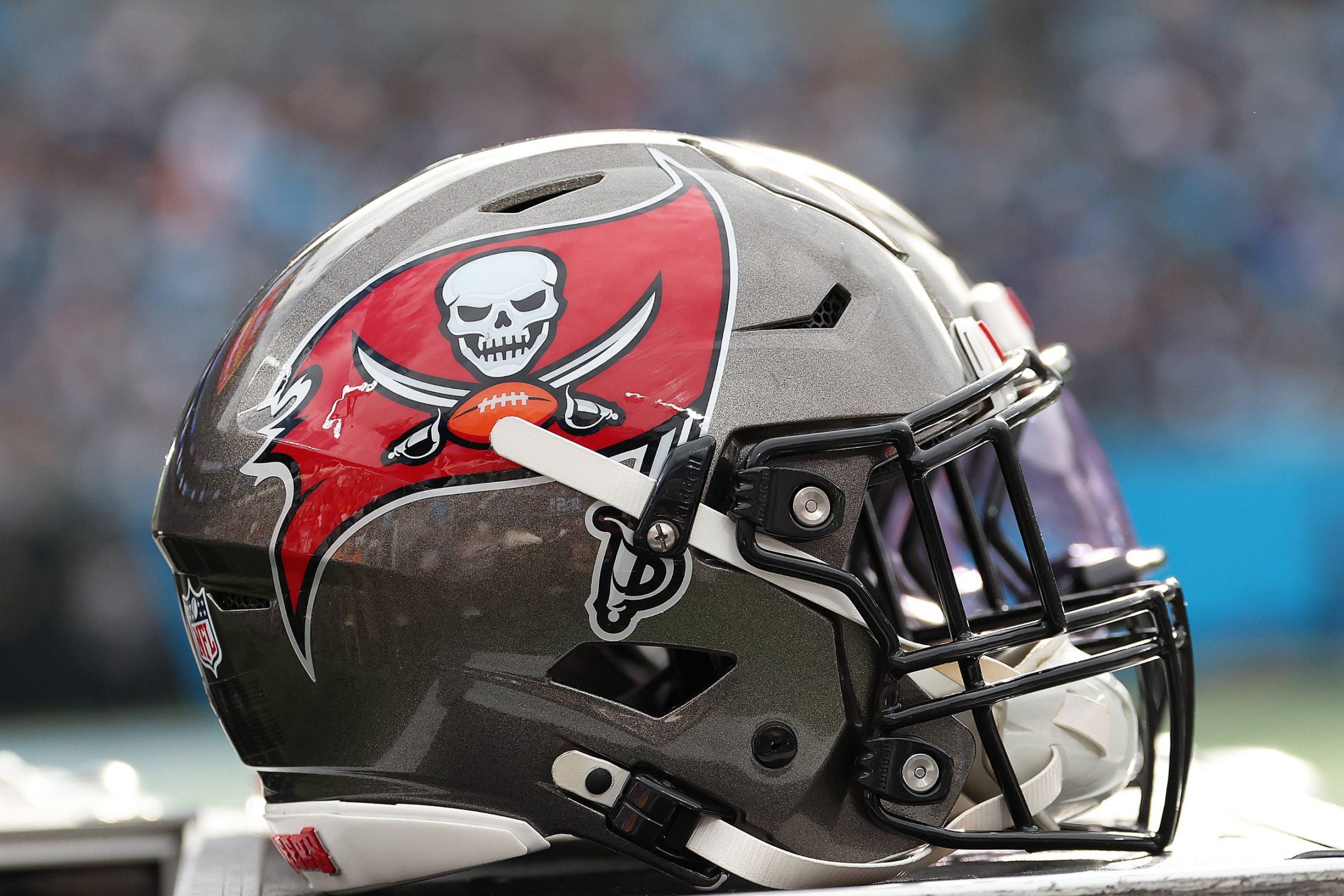 Fünf Trainer mussten gehen - Eine Nahaufnahme eines Footballhelms der Tampa Bay Buccaneers zeigt ein silbernes und rotes Design mit dem ikonischen Piratenflaggen-Logo. Der Helm, der mit Cheftrainer Bowles verbunden ist, ruht auf einer Fläche mit einem unscharfen Stadionhintergrund. Diese Beschreibung wurde mit der FootballR KI automatisch generiert.