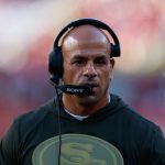 Ein Mann mit Glatze, der dem Titans-Cheftrainer Robert Saleh ähnelt, trägt auf einem Football-Feld ein Headset mit einem Sony-Mikrofon. Er trägt ein grünes Kapuzensweatshirt mit einem goldenen "SF"-Logo. Der unscharfe Hintergrund zeigt ein Stadion und einen Spieler in rot-goldener Kleidung. Diese Beschreibung wurde mit der FootballR KI automatisch generiert.