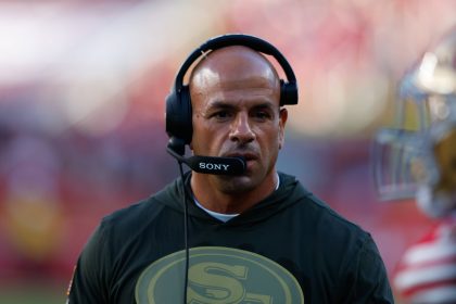 Ein Mann mit Glatze, der dem Titans-Cheftrainer Robert Saleh ähnelt, trägt auf einem Football-Feld ein Headset mit einem Sony-Mikrofon. Er trägt ein grünes Kapuzensweatshirt mit einem goldenen "SF"-Logo. Der unscharfe Hintergrund zeigt ein Stadion und einen Spieler in rot-goldener Kleidung. Diese Beschreibung wurde mit der FootballR KI automatisch generiert.