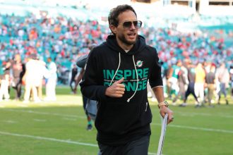 Mike McDaniel - Ein Mann mit Sonnenbrille und schwarzem "Inspire Change"-Kapuzenpulli joggt über ein leuchtend grünes Football-Feld und hält einen Ordner in der Hand. Die verschwommene Menge im Stadion deutet die Energie des Spieltags an - könnte er der nächste Head Coach der Titans sein?. Diese Beschreibung wurde mit der FootballR KI automatisch generiert.