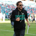 Mike McDaniel - Ein Mann mit Sonnenbrille und schwarzem "Inspire Change"-Kapuzenpulli joggt über ein leuchtend grünes Football-Feld und hält einen Ordner in der Hand. Die verschwommene Menge im Stadion deutet die Energie des Spieltags an - könnte er der nächste Head Coach der Titans sein?. Diese Beschreibung wurde mit der FootballR KI automatisch generiert.