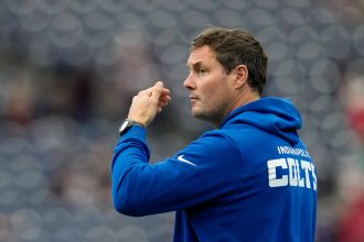 Philip Rivers - Ein Mann, der einen blauen "INDIANAPOLIS COLTS"-Kapuzenpulli trägt, steht im Profil und sieht aus wie Philip Rivers. Er scheint ein Sportereignis zu beobachten oder zu signalisieren, wobei die Sitze im Stadion im Hintergrund verschwimmen. Diese Beschreibung wurde mit der FootballR KI automatisch generiert.