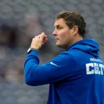 Philip Rivers - Ein Mann, der einen blauen "INDIANAPOLIS COLTS"-Kapuzenpulli trägt, steht im Profil und sieht aus wie Philip Rivers. Er scheint ein Sportereignis zu beobachten oder zu signalisieren, wobei die Sitze im Stadion im Hintergrund verschwimmen. Diese Beschreibung wurde mit der FootballR KI automatisch generiert.