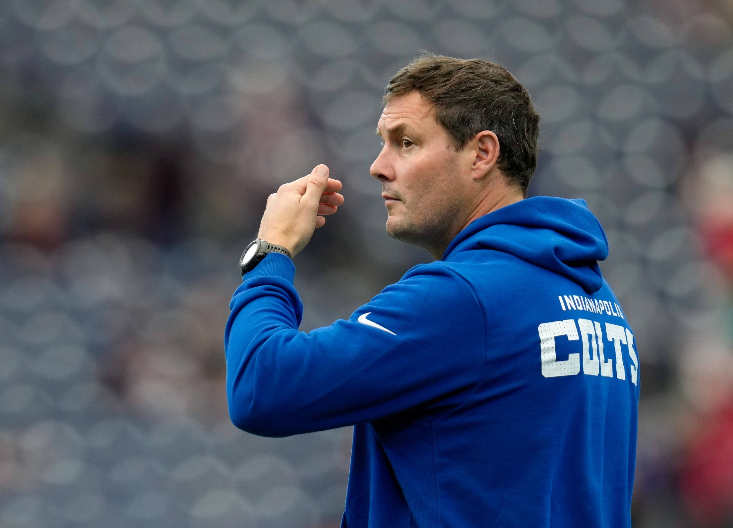 Philip Rivers - Ein Mann, der einen blauen "INDIANAPOLIS COLTS"-Kapuzenpulli trägt, steht im Profil und sieht aus wie Philip Rivers. Er scheint ein Sportereignis zu beobachten oder zu signalisieren, wobei die Sitze im Stadion im Hintergrund verschwimmen. Diese Beschreibung wurde mit der FootballR KI automatisch generiert.